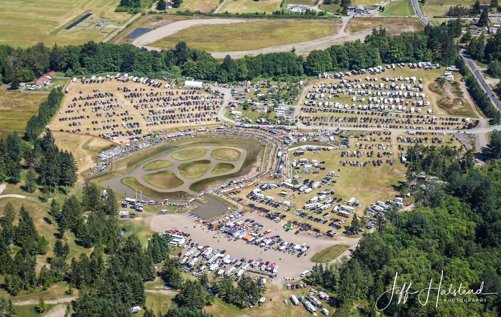 Aerial photo of Extreme Sports Park on a race weekend: the oval sprint boat pond with grassy islands, grandstand along its north edge, and parking and camping filling the fields around it